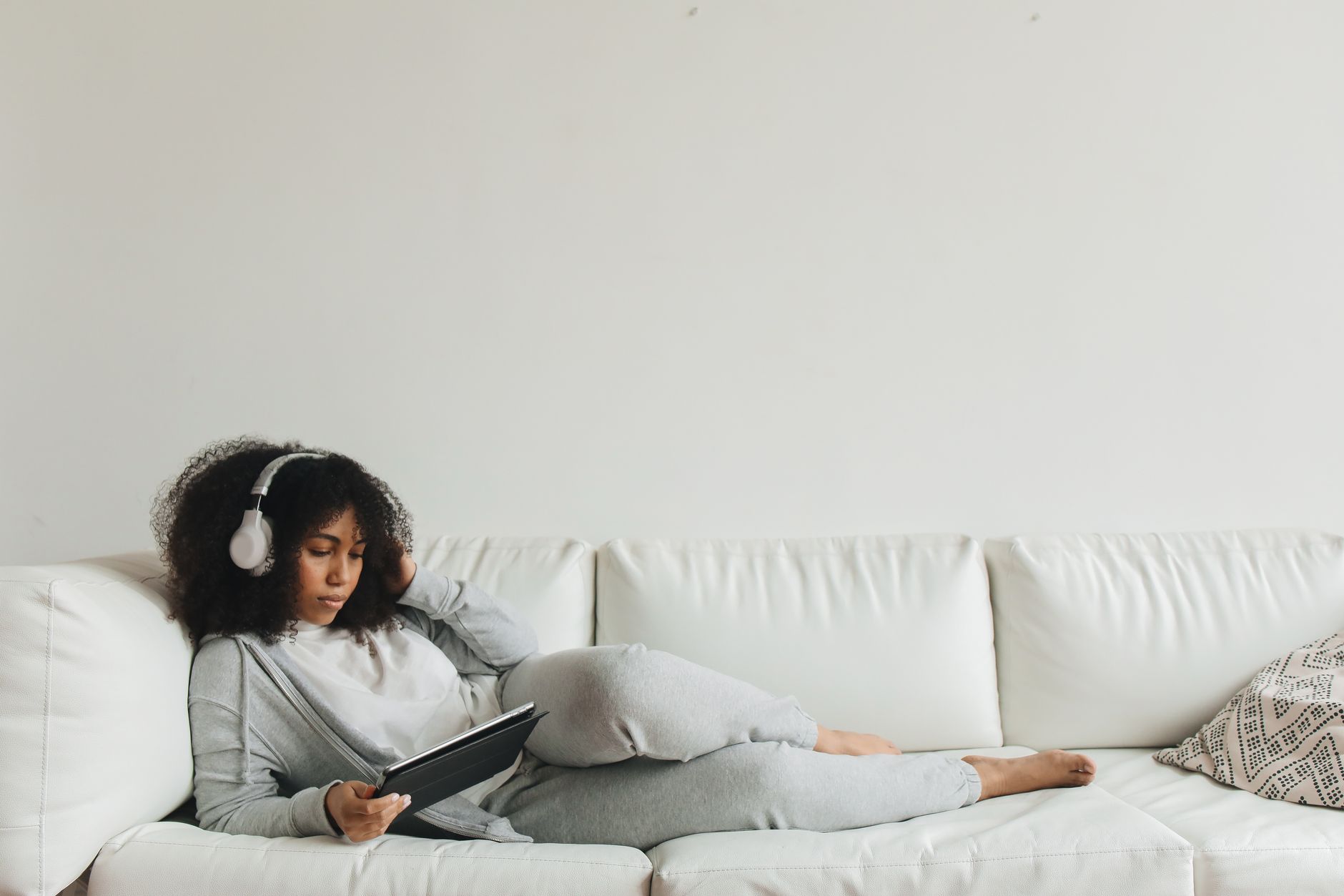 Jeune femme installée sur son canapé, une tablette et un casque à la main, en train de travailler confortablement depuis chez elle