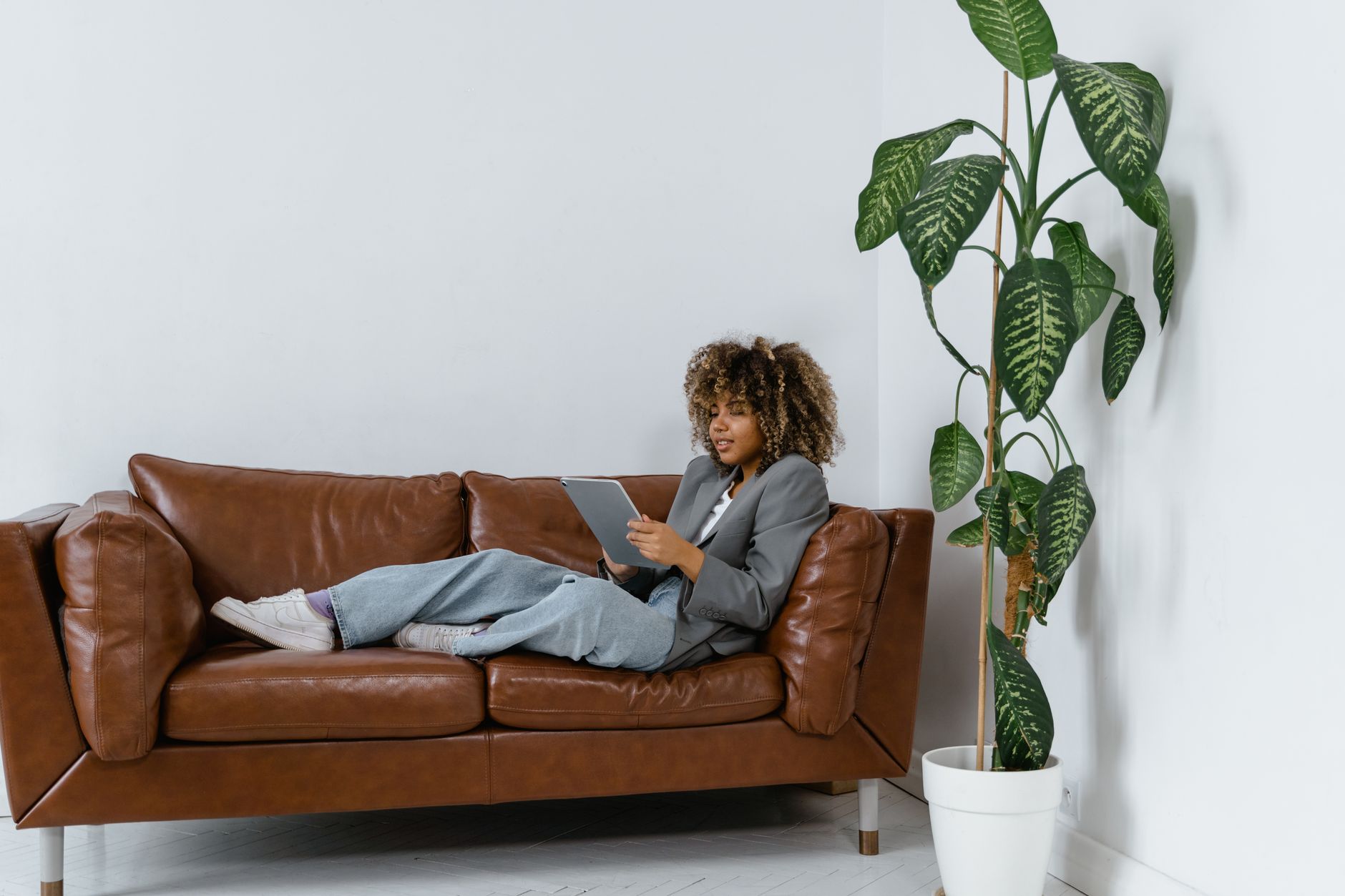 Femme installée confortablement sur un canapé en cuir avec une tablette, à côté d'une plante verte, en train de préparer du contenu